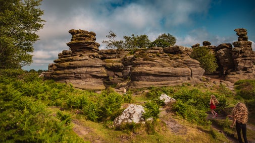 Visitors walk along a narrow path through Brimham Rocks, surrounded by distinctive stacked rock formations, green bracken and moorland vegetation under a partly cloudy sky.